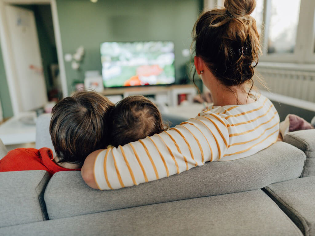 A mother and her two kids watch cartoons on a large screen TV from their couch